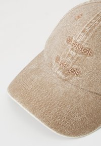 Brown fabric cap with embroidered text. Features a curved brim, textured surface, and detailed stitching along the edges.