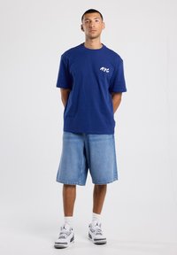 T-paita, jossa on lyhyet hihat, tummansininen ja valkoisella "NYC" -grafiikalla. Yhdistetty väljiin farkkushortseihin ja valkoisiin sneaker-kengiin, joissa on harmaat yksityiskohdat.