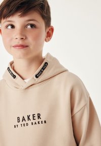 Beige mikina s klokaní kapsou, se černým logem "BAKER". Měkká tkanina, šňůrka s brandovými štítky na výstřihu, uvolněný střih.
