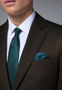 Blazer marron texturé avec une chemise blanche, une cravate verte et un mouchoir de poche teal. Présente des revers crantés et un détail de poche poitrine.