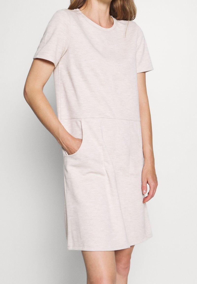 Femme portant une robe beige clair à manches courtes, longueur genou, avec des poches latérales, posant devant un fond clair uni.