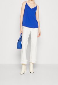 Camisole bleue à bords ondulés, pantalon blanc à jambes larges, bottines blanches et sac à main bleu tissé. Tissu lisse avec une coupe décontractée.