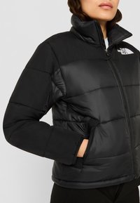 Chaqueta acolchada negra con cuello alto, cierre de cremallera y bolsillos laterales. Presenta paneles acolchados y una textura brillante, destacando el aislamiento.