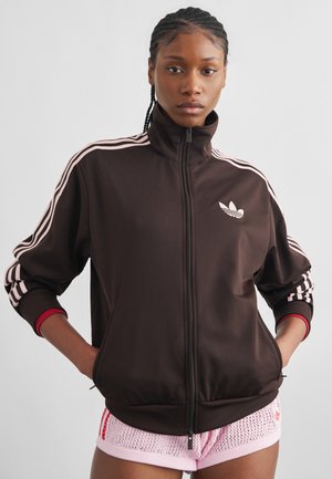 Noor naine punutud juustega, kandes musta lukuga jopet roosade triipudega ja roosaid tekstuuriga lühikesi pükse, seisab käed taskutes.