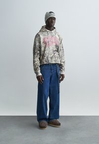 Φούτερ με κουκούλα σε καμουφλάζ σχέδιο και ροζ κείμενο, συνδυασμένο με φαρδιά μπλε τζιν παντελόνια. Αξεσουάρ περιλαμβάνουν γκρι σκουφί και καφέ παπούτσια.