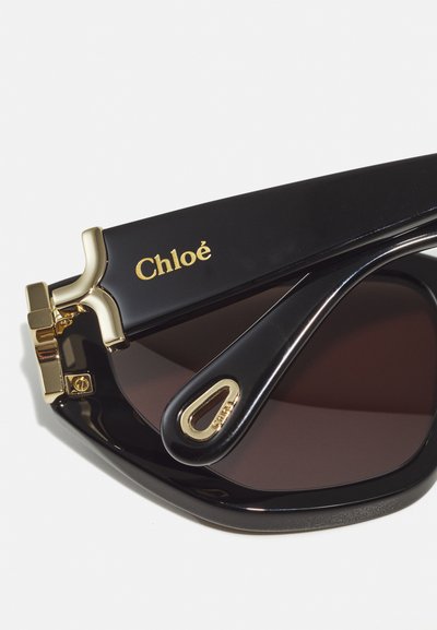 Black sunglasses with brown lenses, featuring a gold logo on the side, textured arms, and a distinct metallic accent near the hinges.