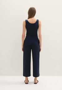 Mono azul marino con un corsé ajustado y piernas anchas, que presenta un escote en la espalda y sandalias tipo deslizamiento. La tela parece suave y ligera.