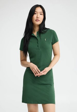 Jonge vrouw met lang zwart haar, gekleed in een donkergroene polo-jurk met korte mouwen, staand tegen een eenvoudige lichte achtergrond.