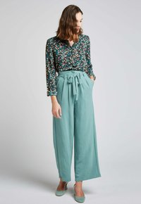 Blouse florale à manches longues et col, associée à un pantalon large taille haute en teal avec une ceinture nouée. Talons en daim bleu.