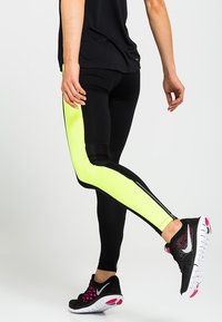 Leggings neri con un pannello laterale giallo neon; presentano un design aderente, una texture liscia e accenti a contrasto. Indossati con scarpe da ginnastica nere.