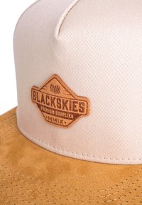 Cappello beige con visiera in suede color beige e una patch in pelle marrone con la scritta "BLACKSKIES PREMIUM SUPPLIES." Tessuto testurizzato, design strutturato.