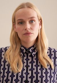 Camicia con colletto in un motivo geometrico viola e crema, dotata di una chiusura frontale con bottoni e una texture di tessuto liscia.