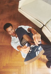 Sötétkék sneaker fehér csíkokkal és fekete fűz�őkkel. Texturált talppal és logós címkével rendelkezik. A viselője keresztbe tett lábbal ülve köti meg a cipőt.