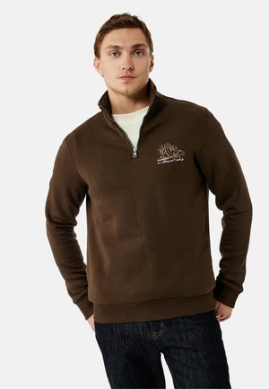 Młody mężczyzna ubrany w brązową bluzę z suwakiem do ćwiartki z grafiką góry i napisem, w połączeniu z ciemnoniebieskimi jeansami, stojący na białym tle.