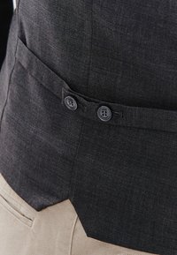 Ciemnoszara, teksturowana kurtka z dwoma czarnymi guzikami z boku, noszona na jasny beżowy strój, prezentująca dopasowany krój.