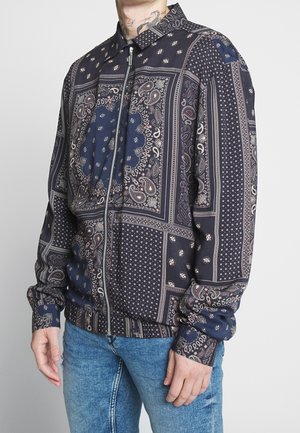 Mężczyzna ubrany w granatową kurtkę zapinaną na zamek błyskawiczny z misternymi wzorami paisley i geometrycznymi, w połączeniu z niebieskimi jeansami, stojący na jednolitym tle.