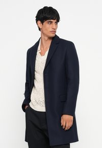 Palton din lână bleumarin cu guler ascuțit și închidere cu un singur buton, asortat cu un pulover tricotat de culoare crem, purtat peste pantaloni negri.