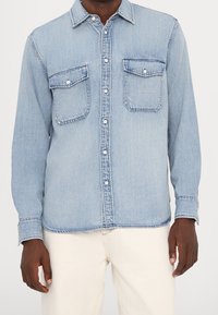 Camisa de mezclilla azul claro con mangas largas, cierre frontal con botones, dos bolsillos en el pecho y detalles visibles de costura.