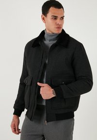 Črn bomberski jopič z umetnim krznom na ovratniku, dvema sprednjima žepoma, pletenimi manšetami in zadrgo, nošen preko sivo rebraste turtleneck majice.