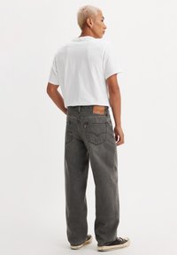 Baggy grey jeans with a faded finish, placed high on the waist, featuring a brown leather patch and back pockets. Paired with a white t-shirt.