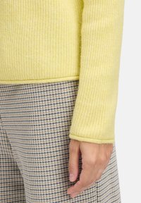 Main reposant sur le côté portant un pull en maille jaune et un pantalon à motifs à carreaux marron sur fond clair.