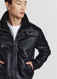 Giovane uomo che indossa una giacca nera con zip e un colletto alto e peloso, con le mani nelle tasche, che guarda di lato su uno sfondo grigio uniforme.