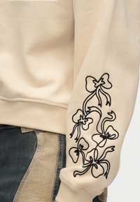 Μανίκι μπεζ φούτερ με μαύρο κεντητό σχέδιο λουλουδάτης κορδέλας σε συνδυασμό με τζιν παντελόνι patchwork που δείχνει ανοιχτά και σκούρα μπλε πάνελ.