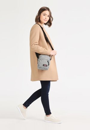 Grå tvärväska med justerbar rem, blixtlåsförsedda frontfickor, tygtextur och logodetalj. Bärs av en person i en beige jacka och jeans.