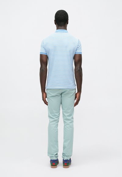 Άνδρας που στέκεται με το πρόσωπο στραμμένο προς τα πίσω, φορώντας ένα polo μπλουζάκι με ουδέτερα γαλάζιες και λευκές ρίγες, πρασινωπό παντελόνι μέντας και μπλε μοκασίνια με πολύχρωμες λεπτομέρειες.
