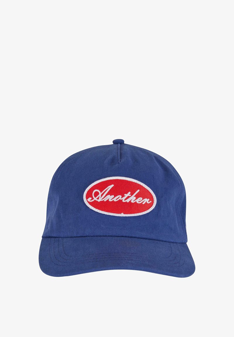 Plava bejzbol kapa napravljena od pamuka s zakrivljenim vizirama. Sadrži crvenu ovalnu zakrpu s bijelim izvezenim tekstom koji glasi "Još jedan."