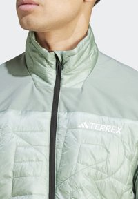 Chaqueta aislante de color verde claro con cuello alto, que cuenta con un cierre frontal de cremallera, textura acolchada y el logo "TERREX" en blanco en el pecho.