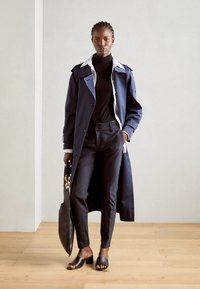 Navy trench coat with epaulets, white collared shirt, black turtleneck, tailored black trousers, and black slide sandals. Minimalist design.