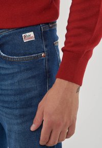 Primo piano di una persona che indossa jeans in denim blu con etichetta "Roy Rogers" e un maglione rosso a maniche lunghe, con la mano rilassata lungo il fianco.