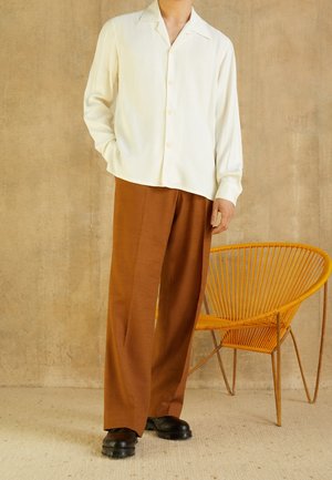 Un homme portant une chemise crème à manches longues boutonnée, un pantalon marron à jambes larges, et des chaussures noires, debout à côté d'une chaise orange tressée.