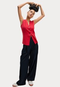 Rotes ärmelloses Top mit tiefem V-Ausschnitt und Knopfdetails, kombiniert mit weiten blauen Hosen und weißen Sneakers. Model hält eine schwarze Tasche.