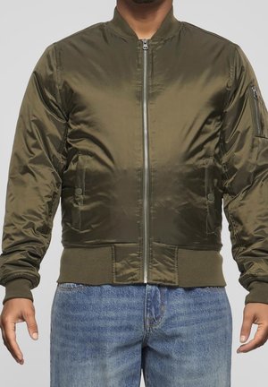 Mężczyzna w oliwkowo-zielonej bomberce z suwakiem z przodu, ściągaczami na mankietach i dole oraz niebieskich dżinsach na jednolitym tle.