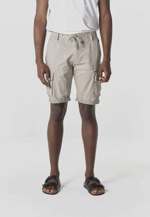 Persona que lleva pantalones cortos cargo beige con dobladillos enrollados, una camiseta blanca y sandalias negras de doble tira, de pie contra un fondo liso.