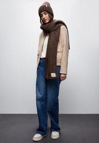 Beige ruskindsjakke, hvid kabelstrik sweater, blå vidde jeans, brun strikket tørklæde og hat, samt beige sneakers med teksturerede detaljer.