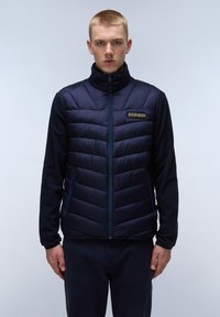 Mörkblå väst med fluffig kviltad design, dragkedja framtill och sidofickor. Har en heltäckande tygkropp och ett märkespatch på bröstet.