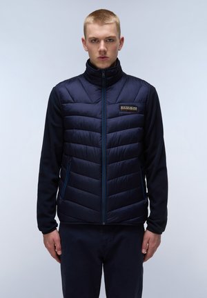 Marineblå vest med puffy quiltet design, lynlås foran og sidelommer. Har en ensfarvet stofoverdel og et mærket patch på brystet.