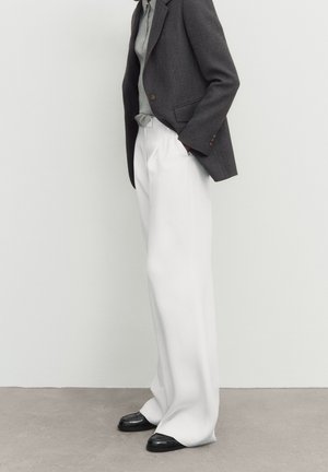 Person wearing a dark gray blazer, light gray top, loose white trousers, and black shoes standing against a plain light background.