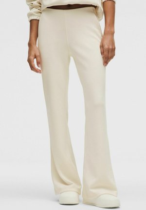 Donna che indossa pantaloni color crema a gamba larga e scarpe slip-on bianche, in piedi contro uno sfondo chiaro e uniforme.