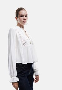 Hvid langærmet bluse med rund halsudskæring, knaplukning og blonderelaterede detaljer. Har en løs pasform og sammenpressede manchetter. Bæres med mørke jeans.