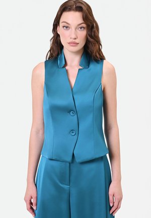 Donna con capelli castani ondulati indossa un gilet in raso turchese senza maniche con colletto e due bottoni, abbinato a pantaloni in raso coordinati.
