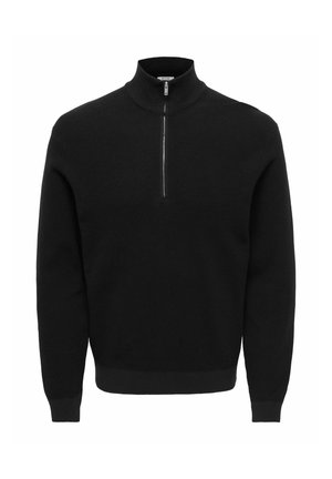 Jeune homme les mains dans les poches, portant une veste et un pantalon beige, une chemise noire en dessous, et des baskets noires, debout sur un fond uni.