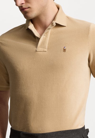 Polo shirt beige a coste con colletto a punte e maniche corte, caratterizzato da un piccolo logo ricamato sul petto. Texture liscia.