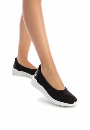 Zapatos negros de punto sin cordones con un acabado texturizado, suela de goma blanca y pequeños acentos metálicos en el talón. Diseñados para la comodidad y la flexibilidad.
