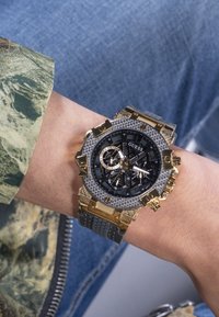 Chronograph-Uhr mit schwarzem Zifferblatt, goldenen Akzenten und strukturiertem grauem Gehäuse. Verfügt über ein Kautschukarmband und mehrere Hilfszifferblätter zur Zeitmessung.