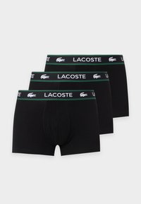 Trois paires de boxers noirs pour hommes avec des ceintures bordées de vert comportant le texte blanc Lacoste et les logos crocodile.