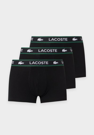 Trois paires de boxers noirs pour hommes avec des ceintures bordées de vert comportant le texte blanc Lacoste et les logos crocodile.